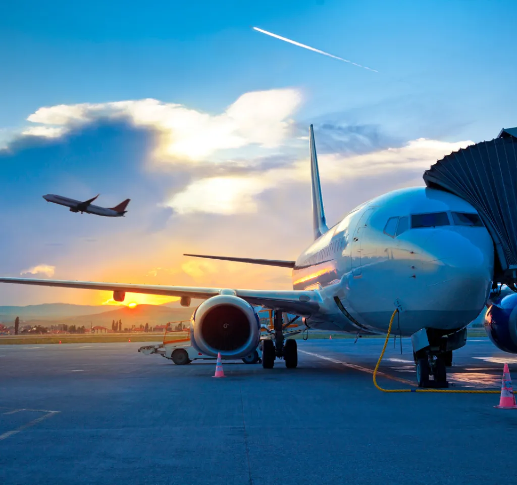 Airplane at gate during sunset with another jet taking off in the colorful sky.