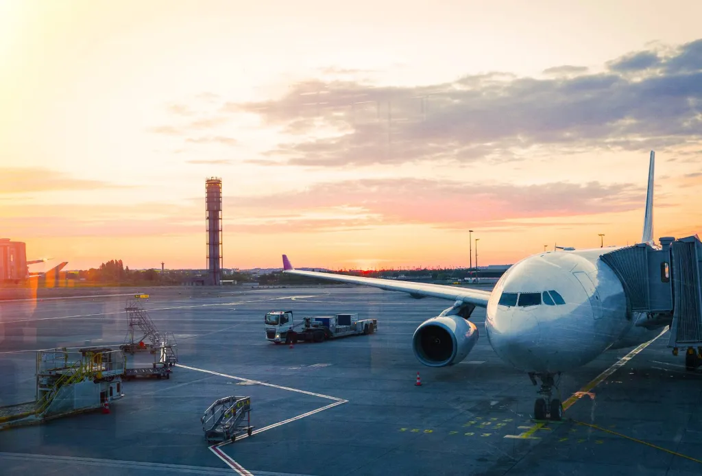 Airplane at airport gate during a colorful sunset, with ground vehicles and equipment nearby.