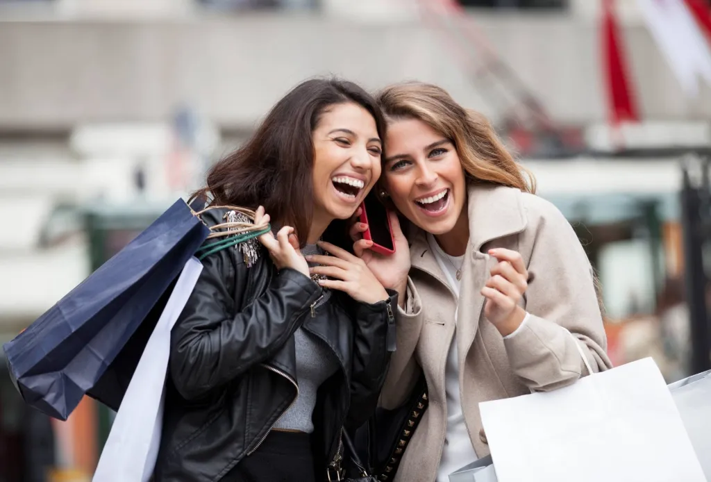 Two women laughing while shopping, holding bags and sharing a phone on a city street.