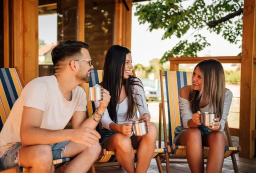 Three friends relaxing on deck chairs, smiling and chatting with mugs in hand.