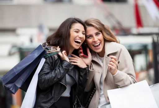 Two women laughing while shopping, holding bags and sharing a phone on a city street.
