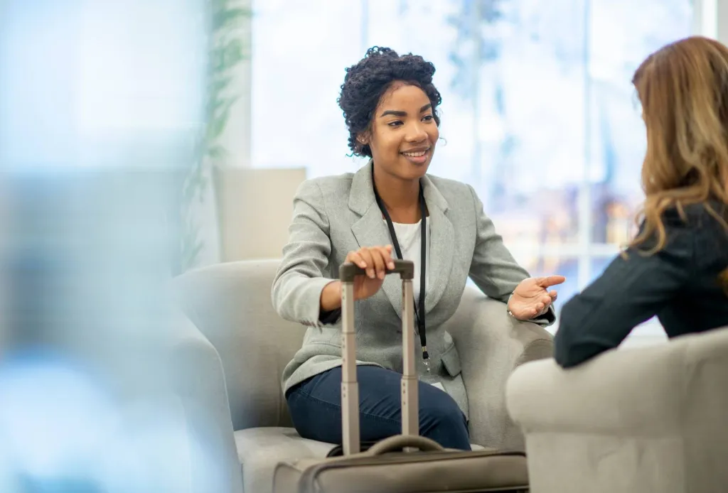 Businesswoman with suitcase talking to a colleague in an airport lounge or office lobby.