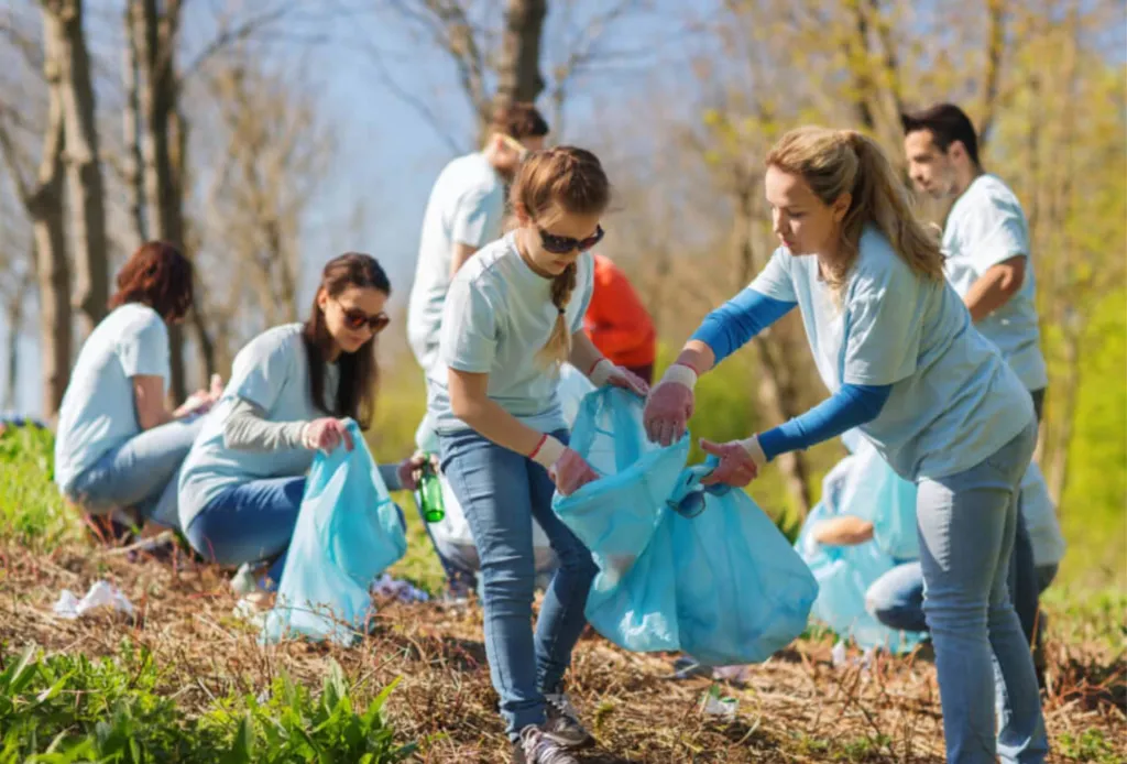 Group of volunteers in light blue shirts picking up litter in a park or forest area.