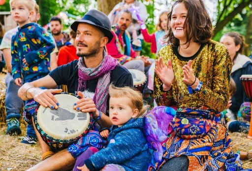 Colorful crowd at outdoor festival with drums, kids, and smiling adults in vibrant clothing.