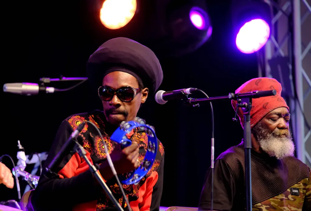 Two reggae musicians performing on stage under colorful lights, one with a tambourine.