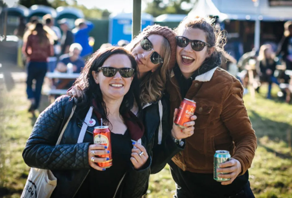 Three friends in sunglasses smiling and holding drinks at an outdoor music festival.