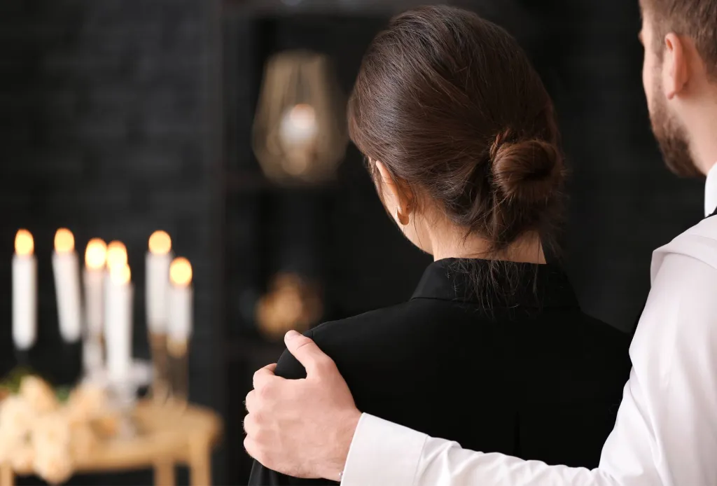 Man comforting woman at a funeral, with lit candles and dark background in soft focus.