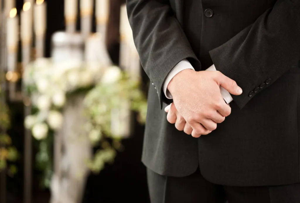 Man in dark suit clasping hands at funeral, with flowers and candles blurred in background.