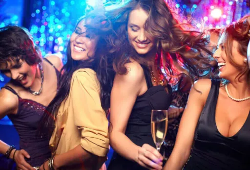 Group of women dancing and smiling in a nightclub with colorful lights and champagne.