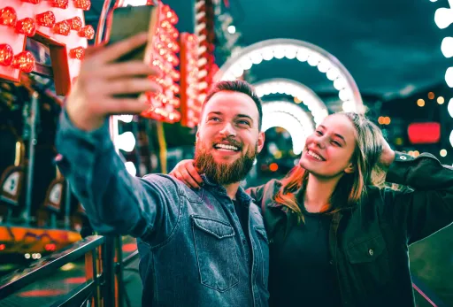 Smiling couple taking a selfie at a brightly lit theme park at night.