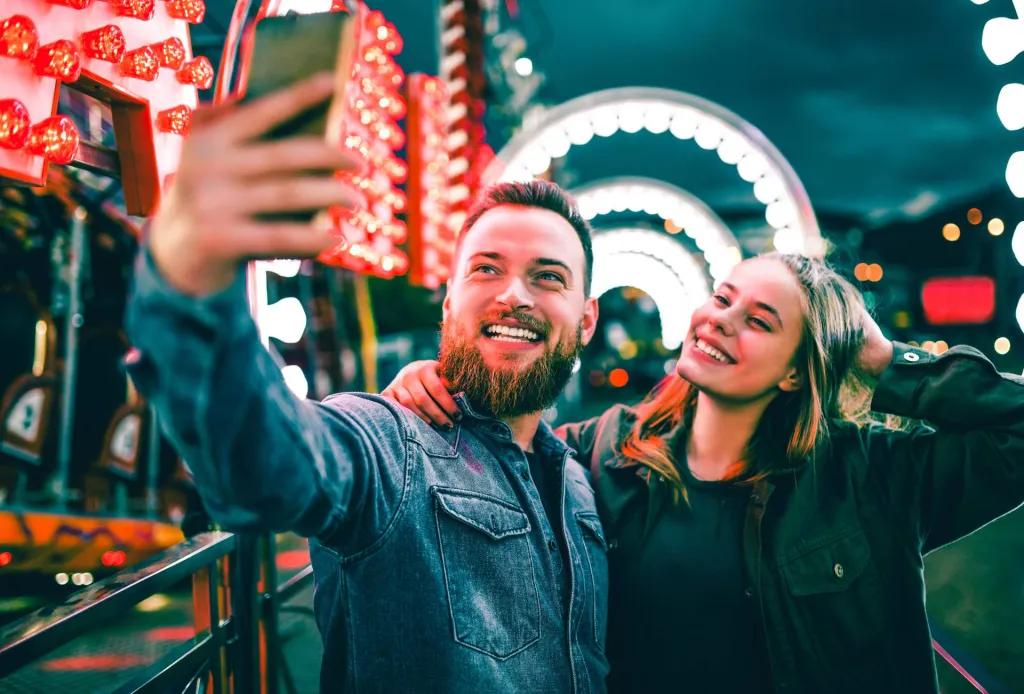 Smiling couple taking a selfie at a brightly lit theme park at night.