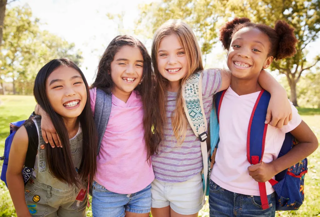 Four young girls with backpacks smiling and hugging outdoors on a sunny day.