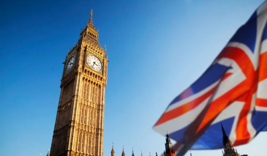 Big Ben clock tower with a waving Union Jack flag in the foreground under a clear blue sky.