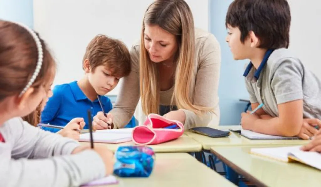 Teacher helping young students with their work at a classroom table.