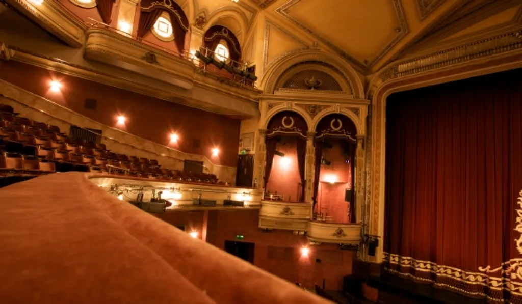 Interior of a grand, historic theatre with ornate balconies and red velvet stage curtain.