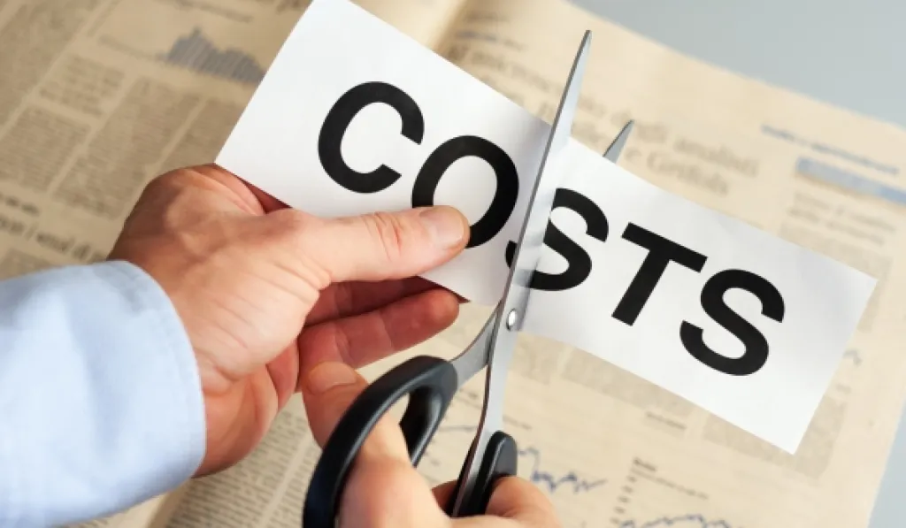 Person cutting a paper labeled COSTS with scissors, symbolizing cost reduction.