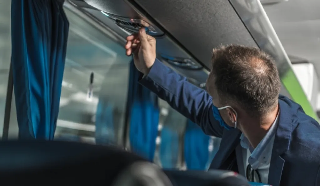 Man adjusting air vent on coach bus while seated, wearing face mask and business attire.