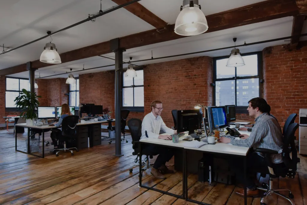 Modern open-plan office with exposed brick walls and employees working at computer desks.